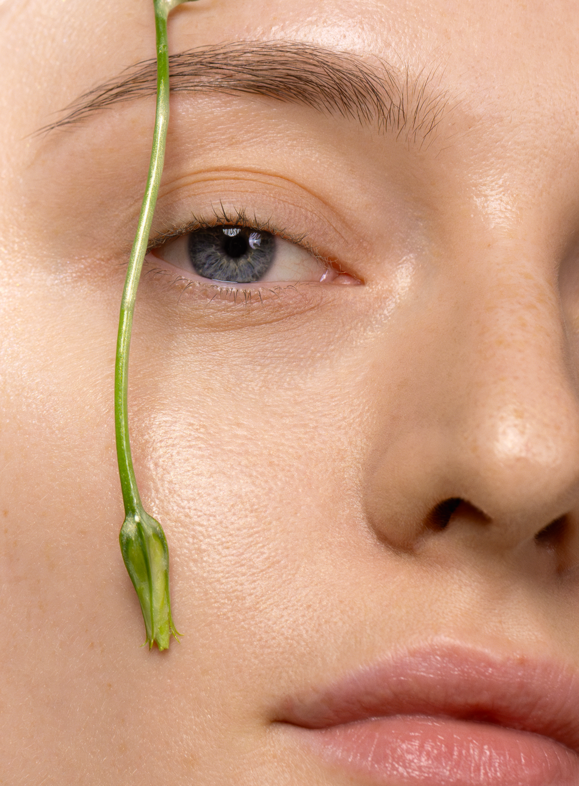 Close-up of a woman’s radiant skin with a green plant stem, symbolizing hydration and natural glow from MÁDARA Hydra Glow Hyaluron Peptide Serum